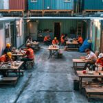 travailleurs-se-detendant-dans-la-cantine-modulaire-dun-chantier-assis-a-des-tables-communes-entourees-dunites-prefabriquees-empilees-avec-boites-repas-tasses-de-cafe-et-casques-poses-eclaira