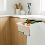 Quels rangements intégrés choisir pour trier les épluchures en cuisine moderne ? Cuisine moderne lumineuse avec façades en chêne clair et blanc mat, tiroir ouvert contenant un petit bac amovible rempli d’épluchures de légumes.