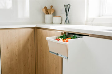 Cuisine moderne lumineuse avec façades en chêne clair et blanc mat, tiroir ouvert contenant un petit bac amovible rempli d’épluchures de légumes.
